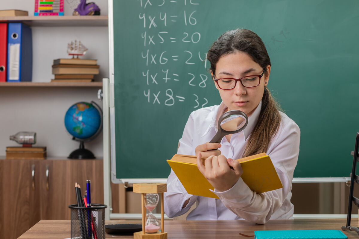 女人年轻的女老师戴着眼镜拿着书通过放大镜看书页自信书桌眼镜