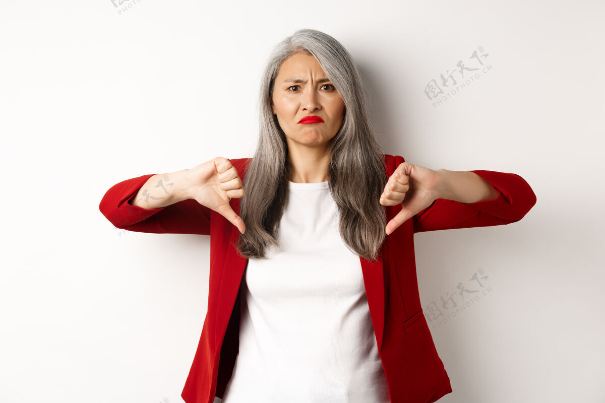 女性穿着红色外套的失望的老妇人竖起大拇指 做鬼脸 不喜欢也不赞成 站在白色的背景下崇拜专业人士批准