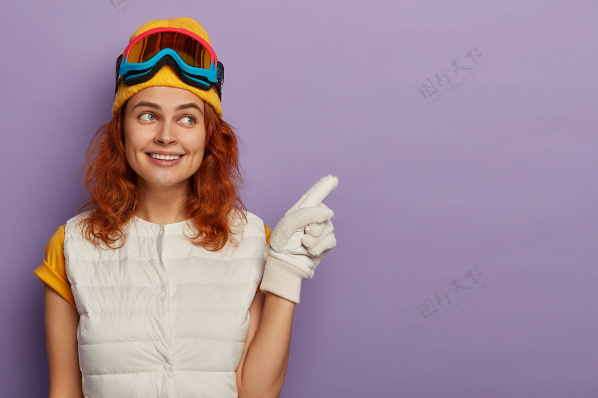 空白迷人的红发女人戴着暖和的帽子 滑雪镜和白色套头衫滑雪者指点青少年