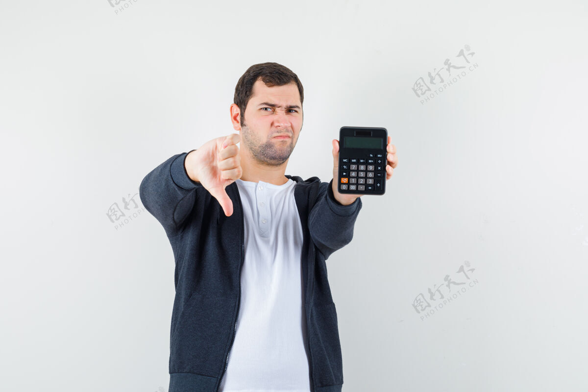 计算器年轻人拿着计算器 大拇指朝下 穿着白色t恤和黑色前拉链连帽衫 看起来很不高兴 正面视图拿着拇指T恤