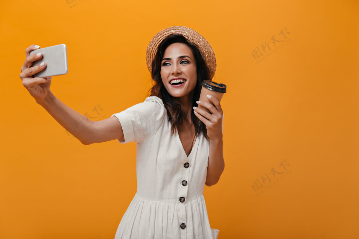 圆点头戴草帽 身穿白色连衣裙的女士在智能手机上自拍 手里拿着一杯茶身着轻装的成年女士手里拿着咖啡拍照划船咖啡年轻