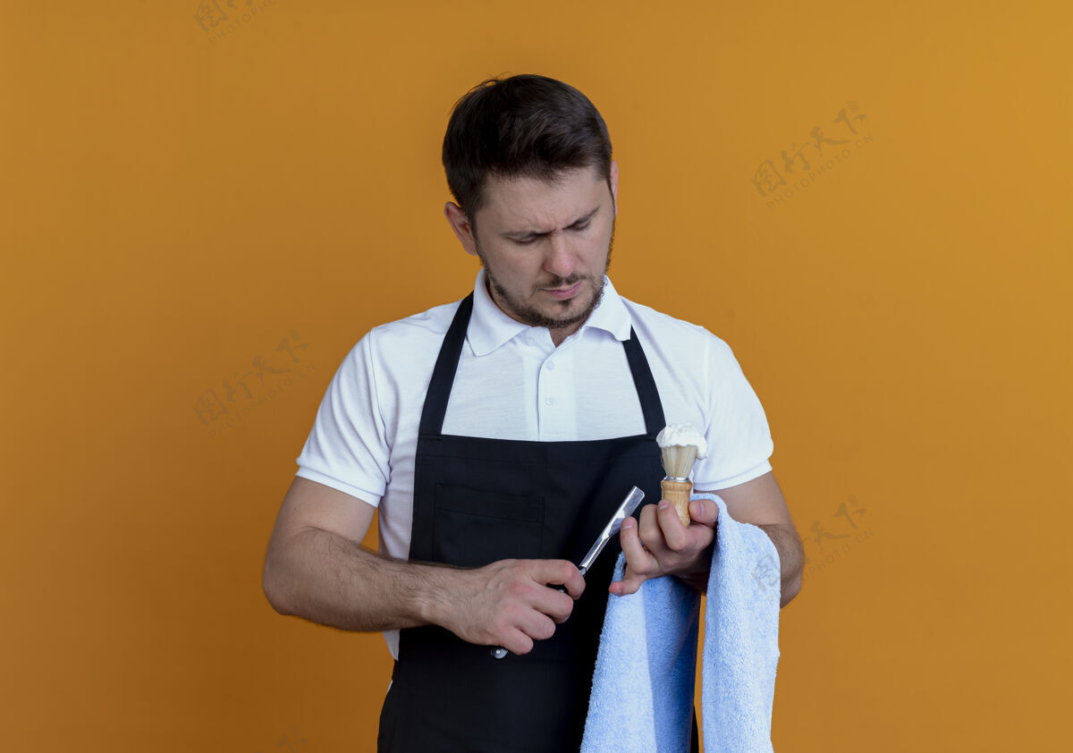 剃须刀围裙上的理发师手拿毛巾 手里拿着泡沫剃须刷和剃须刀 严肃的脸站在橙色的墙上看着它理发师男人围裙