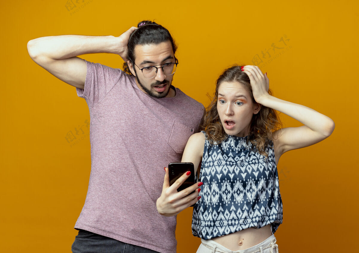 夫妻一对年轻夫妇拿着智能手机站在橙色的墙上 看上去既惊讶又困惑年轻站起来困惑