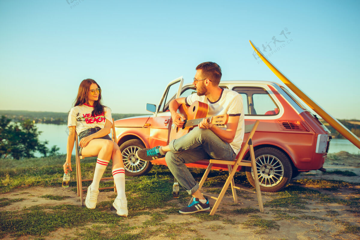 假期一对情侣坐在沙滩上休息 在河边的夏日里弹吉他黄昏自然冲浪板