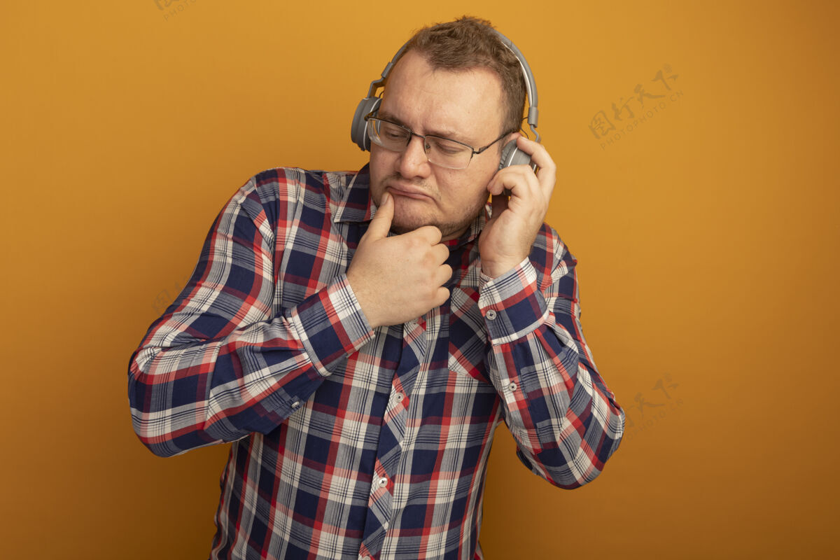 检查一个戴着眼镜 穿着格子衬衫 戴着耳机 闭着眼睛的男人站在橙色的墙上 困惑不解站起来男人眼镜