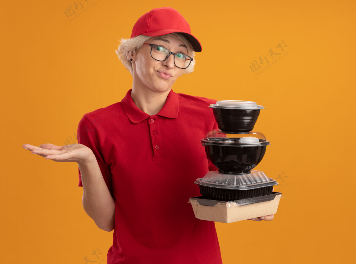 迷糊年轻的送货员身穿红色制服 戴着眼镜 手里拿着一叠食品包 一副迷茫的样子 抬起胳膊站在橙色的墙上女人叠着戴着