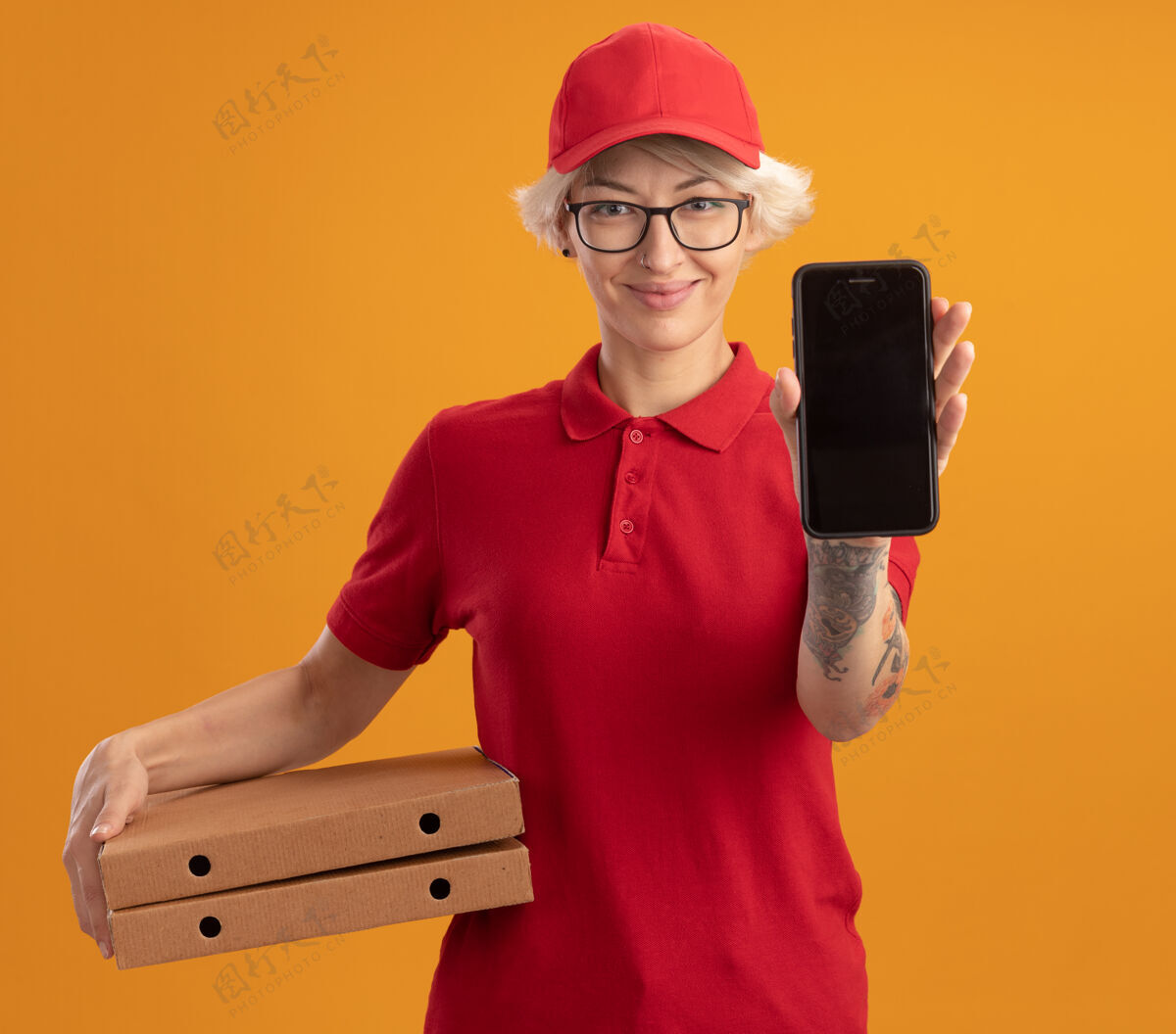 手机身穿红色制服 戴着眼镜 手持比萨饼盒的年轻送货员站在橙色的墙上 脸上洋溢着笑容披萨微笑女士
