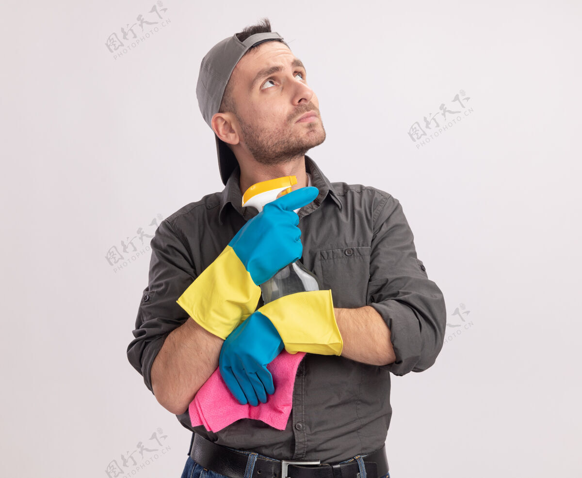 喷雾年轻的清洁工穿着休闲服 戴着橡胶手套 手里拿着喷雾瓶和抹布 站在白色的墙上困惑地看着一边磨损清洁破布