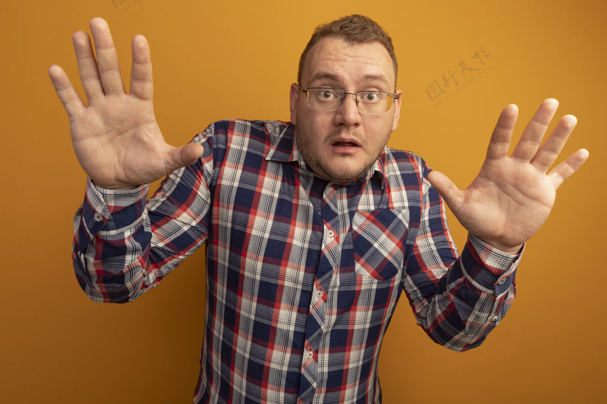 眼镜一个戴着眼镜 穿着格子衬衫的男人站在橙色的墙上 举手投降站着手担心
