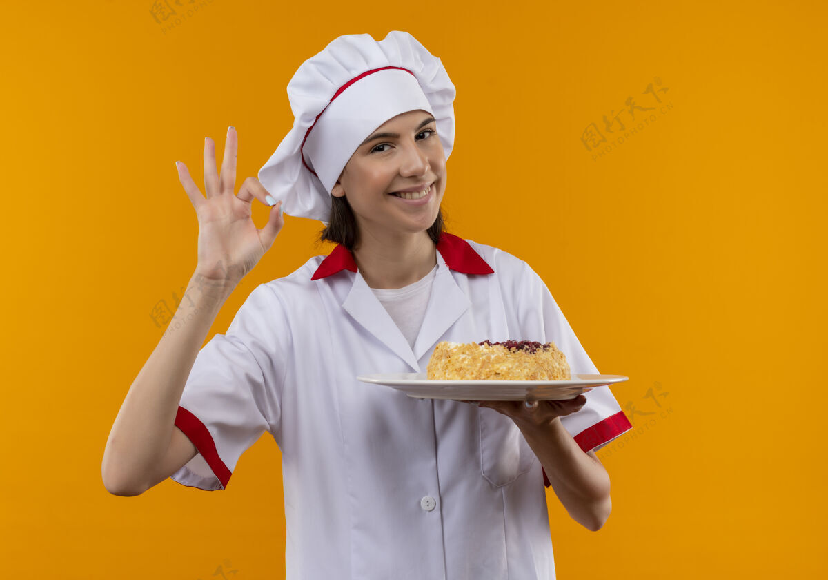烹饪身着厨师制服 面带微笑的白人年轻厨师女孩拿着盘子上的蛋糕 在橙色的空白处用手势表示“ok”的手势年轻制服微笑