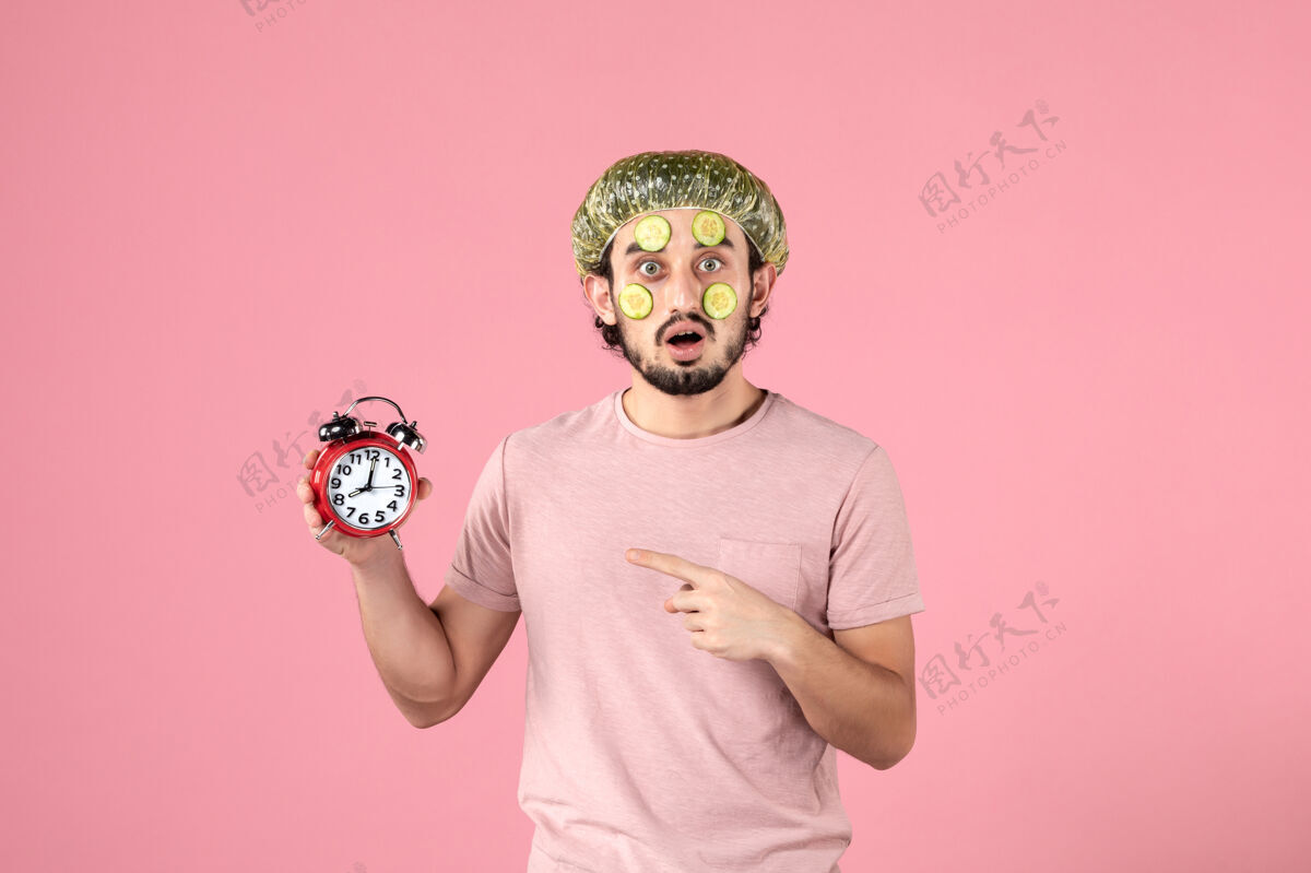 光滑正面图：戴着面具的年轻男性手持粉色背景的时钟面膜视图美容