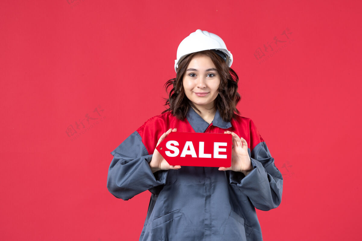 吸引正面是穿着制服 戴着安全帽的自信女工的近景 在孤立的红墙上展示着销售图标前面显示漂亮