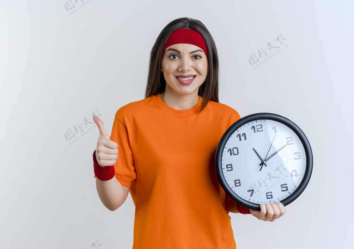 显示微笑着的年轻运动女性戴着头带和腕带拿着时钟 大拇指向上竖立在白色的墙上 留着复制空间女人穿运动