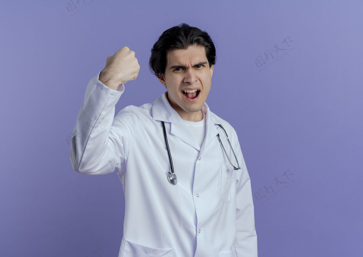 年轻自信的年轻男医生穿着医用长袍和听诊器做着强烈的手势 隔离在紫色的墙上 留着复印空间手势医学强壮