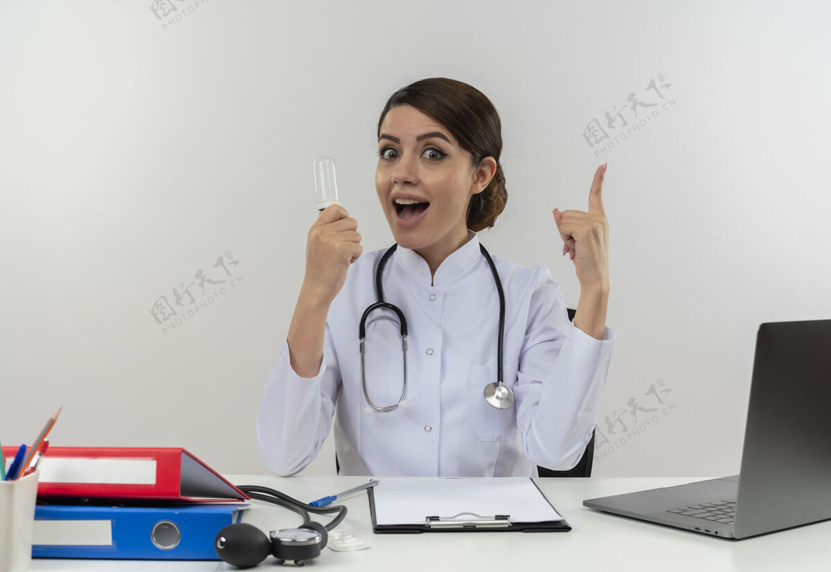 年轻人年轻的女医生穿着医用长袍和听诊器坐在办公桌旁 手里拿着医疗工具和笔记本电脑 手里拿着灯泡 竖起手指隔离在白色的墙上灯光工具办公桌