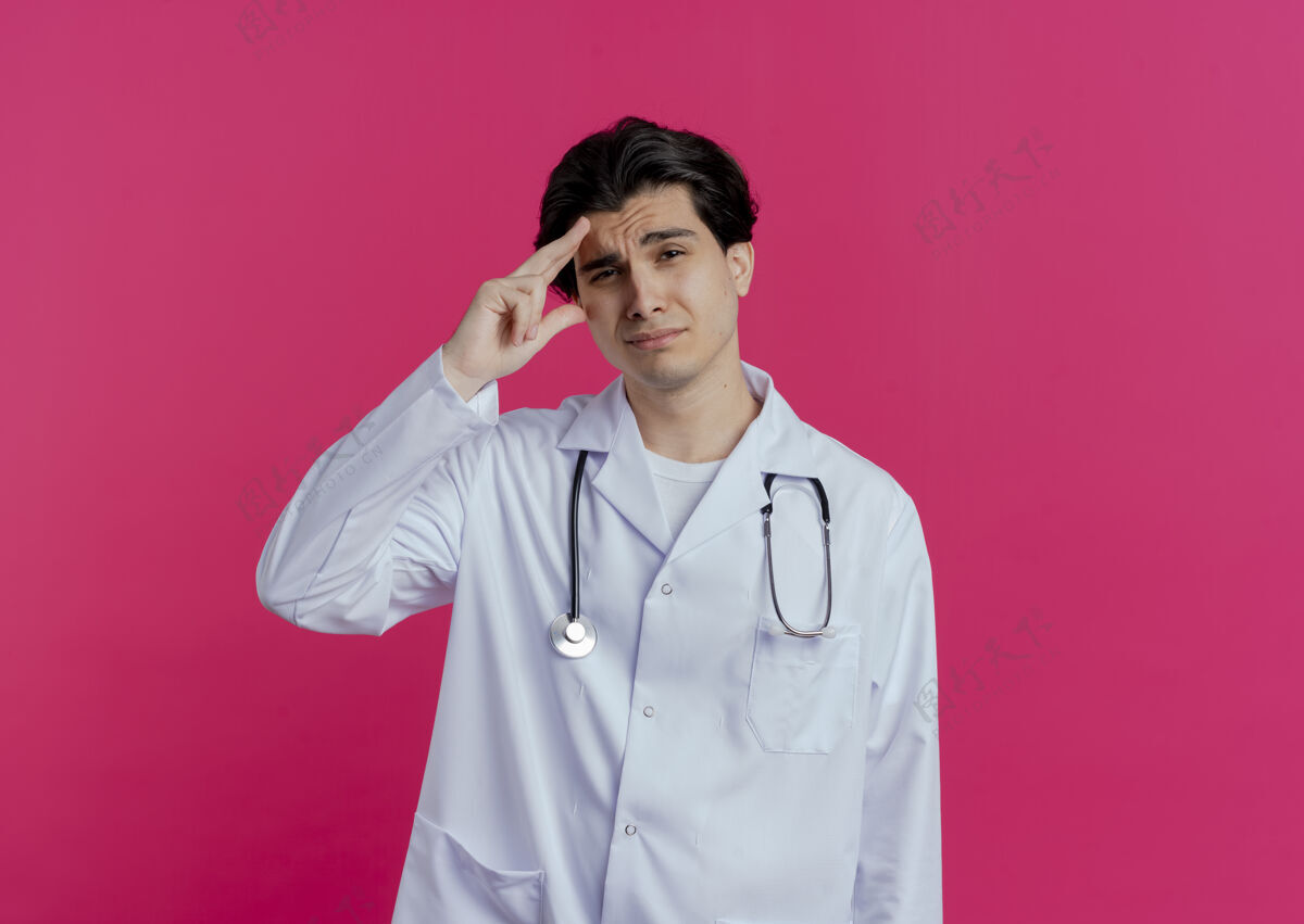 年轻可疑的年轻男医生穿着医用长袍 用听诊器触摸隔离在粉红色墙上的前额 留有复印空间摸听诊器怀疑