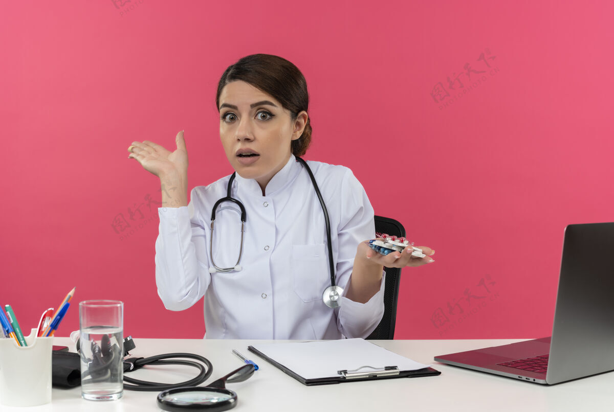 显示年轻的女医生穿着医用长袍和听诊器坐在办公桌旁 手里拿着医疗工具和手提电脑 手里拿着药品 在粉红色的墙上显示出一只孤立的空手女空办公桌