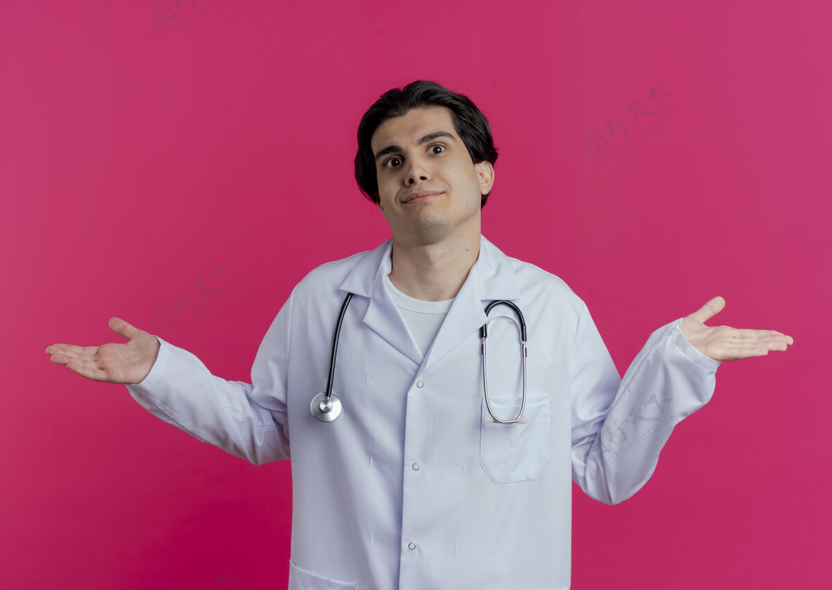 空困惑的年轻男医生穿着医用长袍和听诊器 两手空空的隔离在粉红色的墙上医疗医生穿