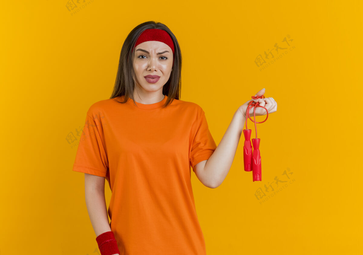 跳跃戴着头带和手环拿着跳绳的未被释放的年轻运动女性被隔离在橙色的墙上 有复制空间看头带绳子