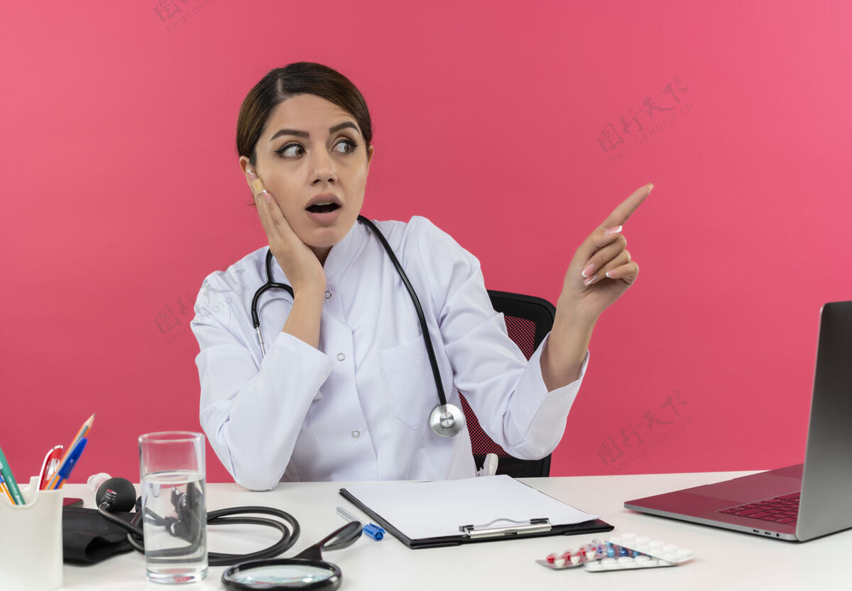 保持惊讶的年轻女医生穿着医用长袍和听诊器坐在办公桌旁 手里拿着医疗工具和笔记本电脑 一边看着一边 手放在脸上 隔离在粉红色的墙上脸穿长袍