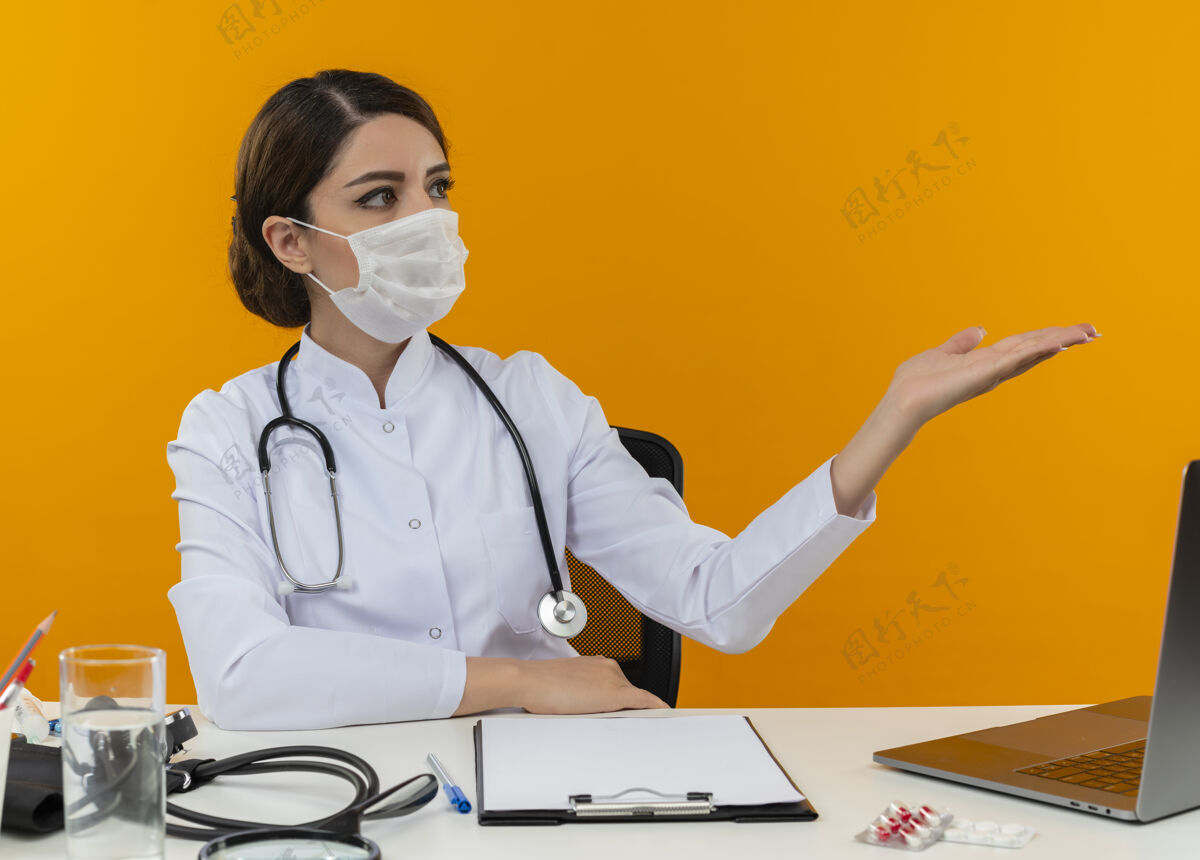 面罩年轻的女医生穿着医用长袍 戴着听诊器 戴着口罩 坐在办公桌旁 拿着医疗工具和笔记本电脑 一边看着黄色墙壁上孤立的空手壁板办公桌医生