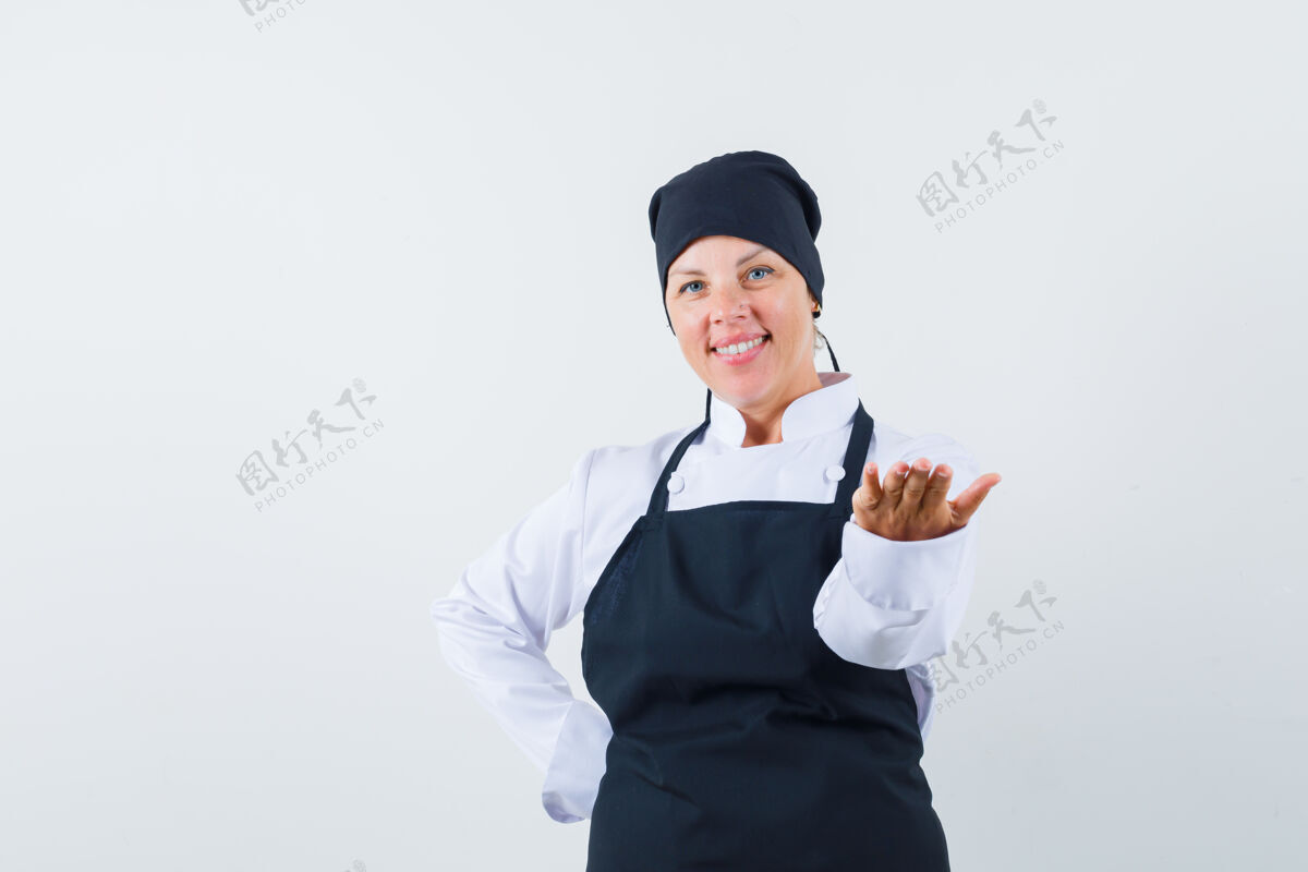 健康一个金发女人把手伸向前面拿东西 另一只手放在腰上 穿着黑色厨师制服 看起来很漂亮可爱自然黑色