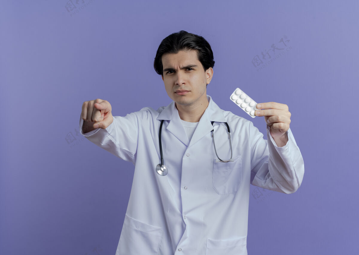 看皱眉的年轻男医生穿着医用长袍 听诊器显示一包药片 看着并指着紫色墙上的复制空间孤立皱眉显示年轻