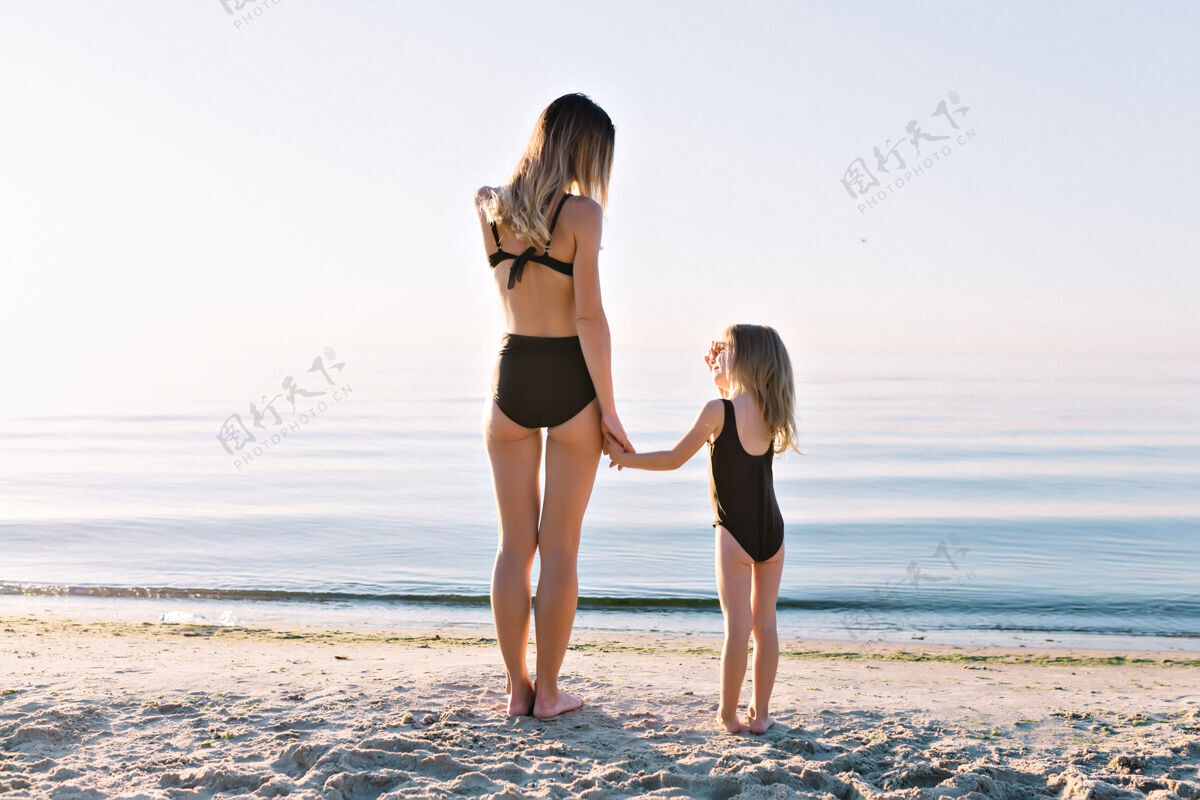 户外年轻漂亮的母亲和穿着黑色泳衣的小女儿在夏日沙滩上孩子小快乐