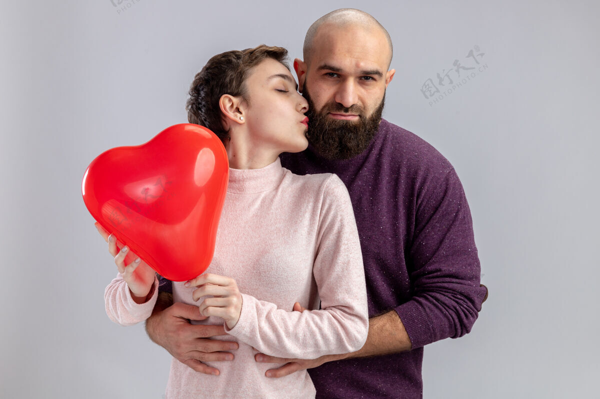 休闲一对穿着休闲服的年轻夫妇手持心形气球的女人站在白色的墙上亲吻着她快乐的男友庆祝情人节形状年轻衣服