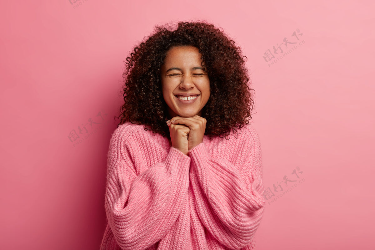 黑发快乐的女孩 蓬松的头发 双手合拢在下巴下 快乐地实现目标 闭着眼睛 微笑着 在粉红色的工作室墙上 被隔离开来Swoosh积极发型