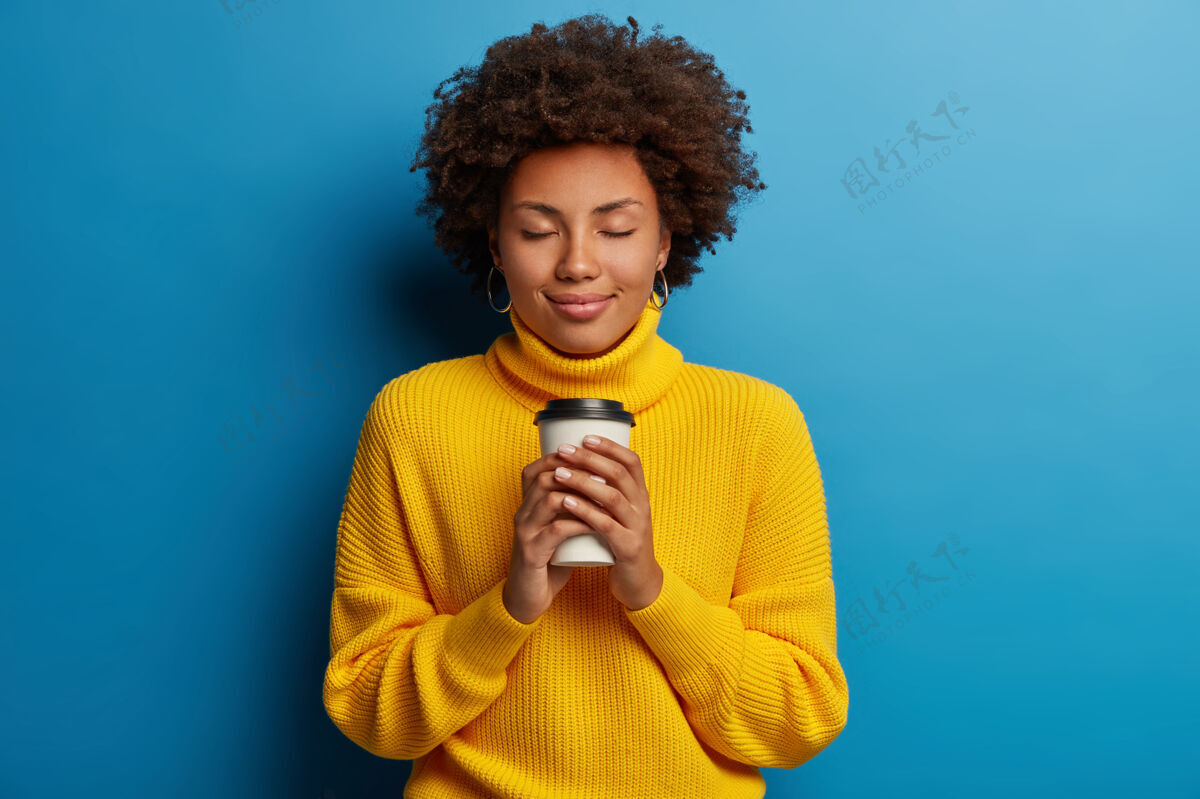 女性满足的年轻非洲女人喜欢喝饮料 拿着外卖咖啡 闭上眼睛 穿着黄色毛衣 隔离在蓝色背景下人肖像微笑