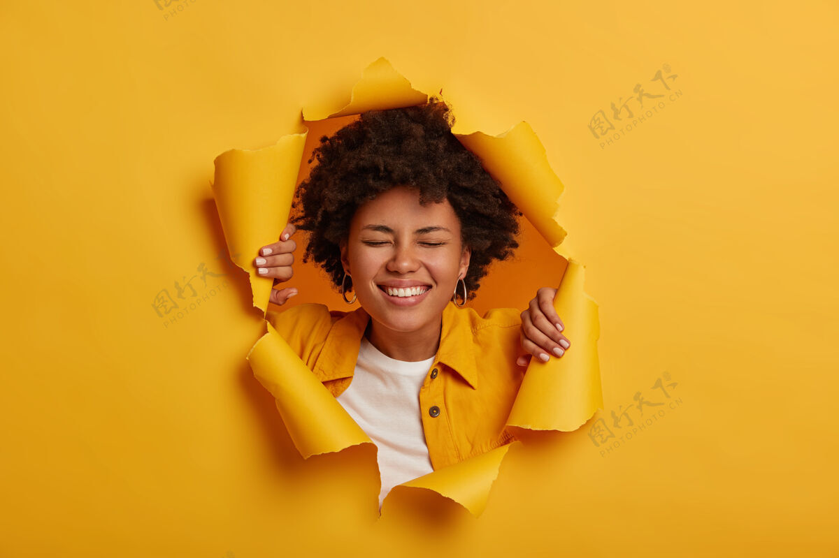 年轻幸运的喜出望外的非裔美国妇女笑容灿烂 心情悠闲 身着时髦服装在黄色的纸背景下摆出姿势人兴趣撕破