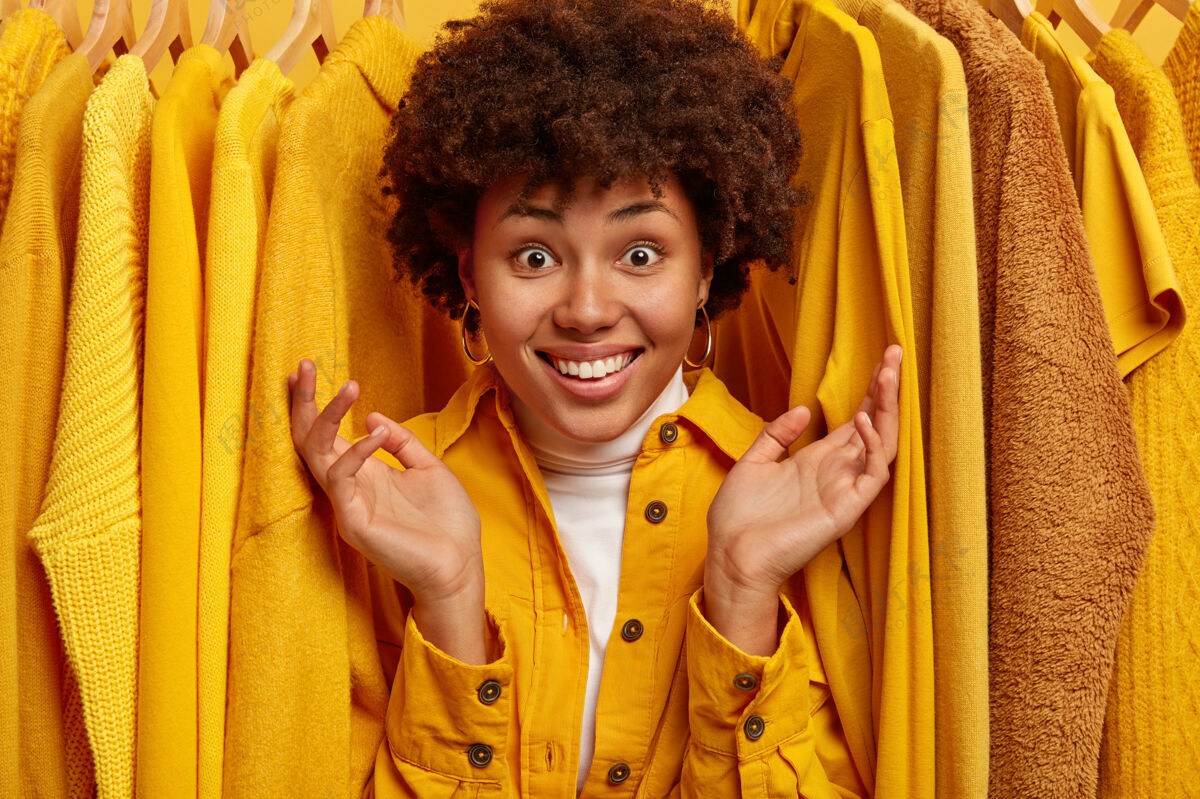 阴性快乐的非洲女人 带着牙齿般的微笑 抬起手掌 展示店里各式各样的衣服 透过衣架上的黄色衣服看正片ShopNew