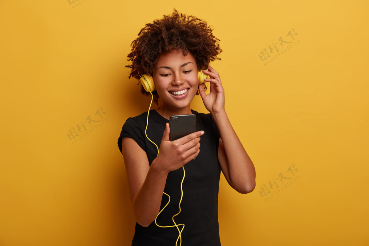 耳机一个活泼活力的女人的肖像 卷发 观看有趣的视频 戴着耳机连接到智能手机隔离在黄色背景上高兴感觉女人