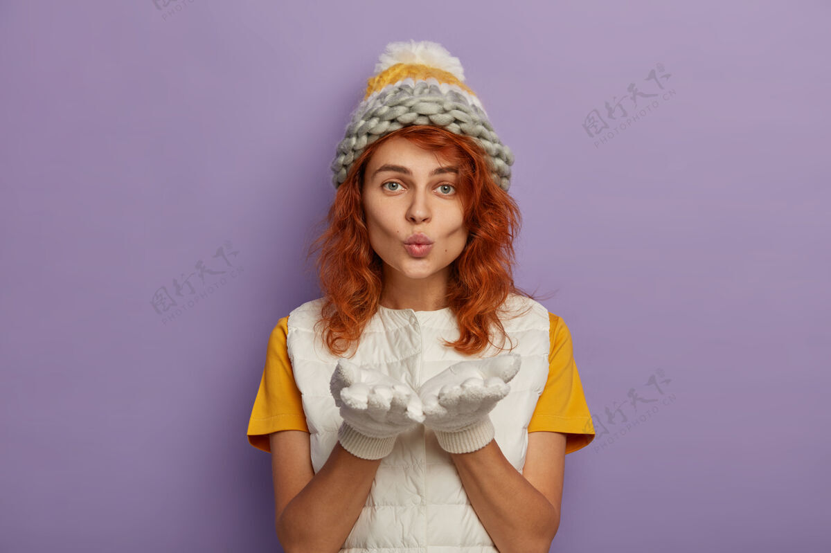 女性红发女人对着镜头吹 手掌向前 戴着针织帽子和白色背心 享受冬天的时光肖像亲吻横