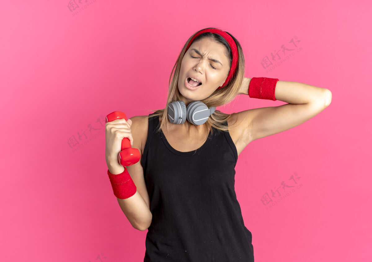 健身身穿黑色运动服 头戴红色头带 手持哑铃的年轻健身女孩 因粉色而显得既不高兴又沮丧年轻抱头带