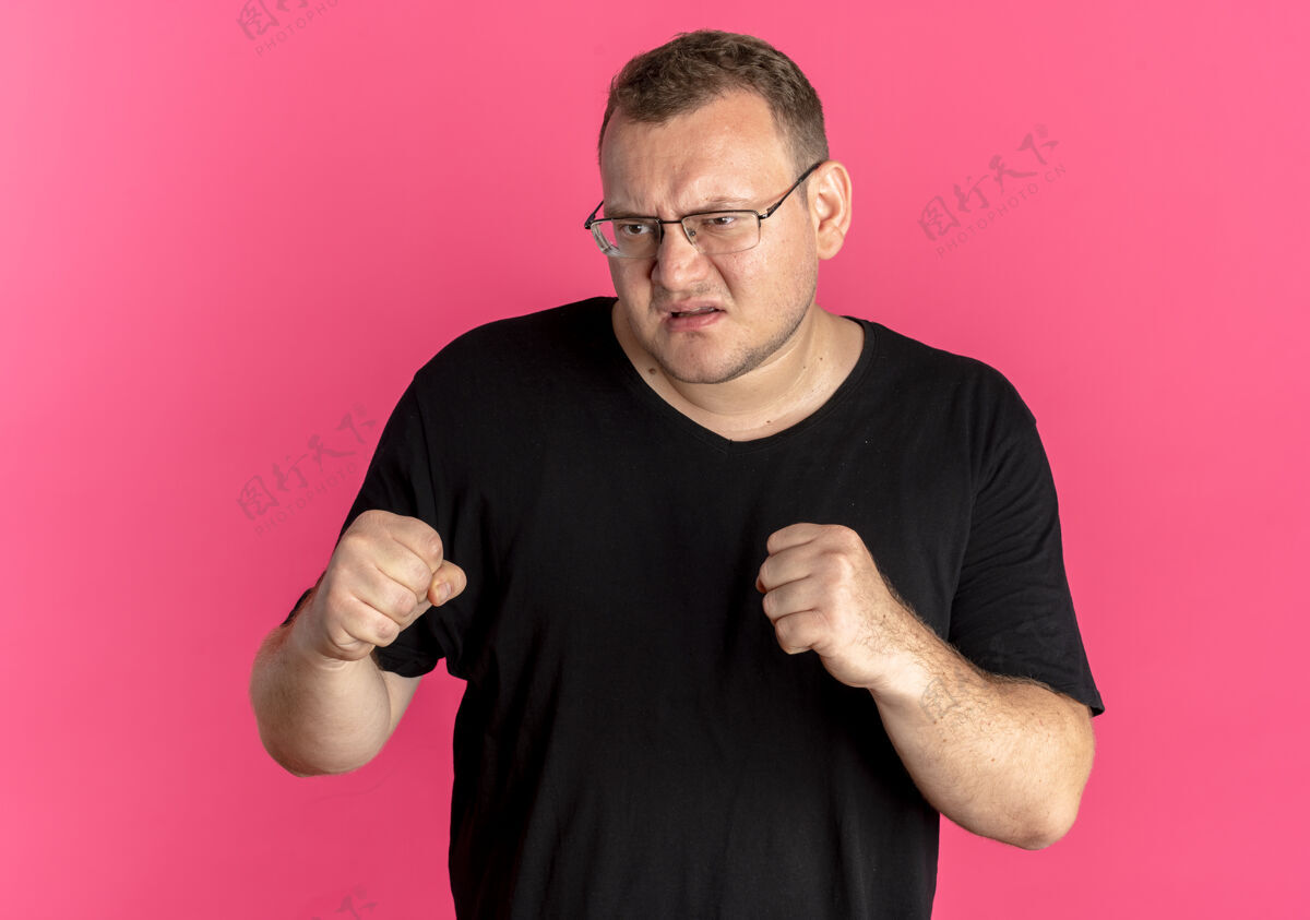 超重戴眼镜的超重男子身穿黑色t恤握紧拳头看着愤怒的脸超过粉红色脸穿粉色
