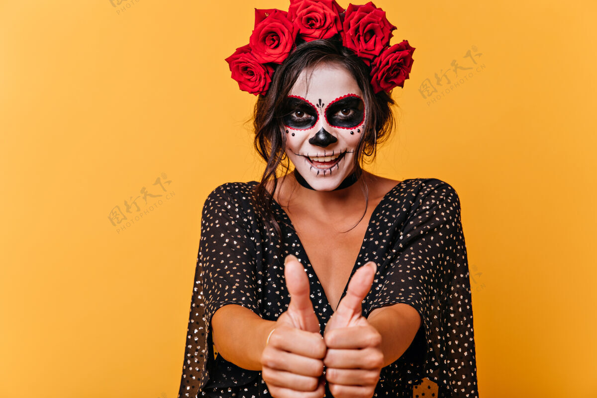 油漆狡猾的棕色眼睛的黑发女子竖起大拇指的肖像戴着玫瑰冠的女孩 以骷髅微笑的形式展现面部艺术头骨深色女性