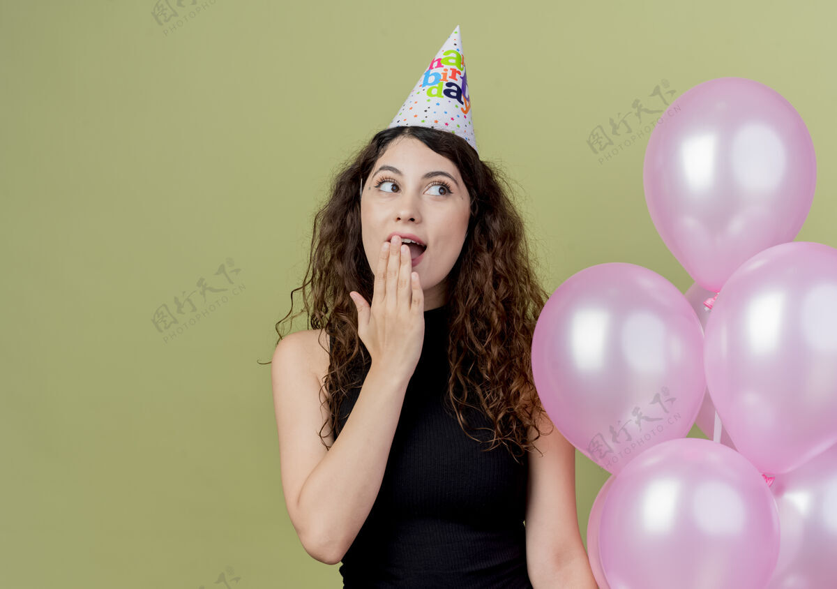 头发戴着节日帽的卷发美女手持气球 一边看着灯光下的惊喜和快乐光气球帽子