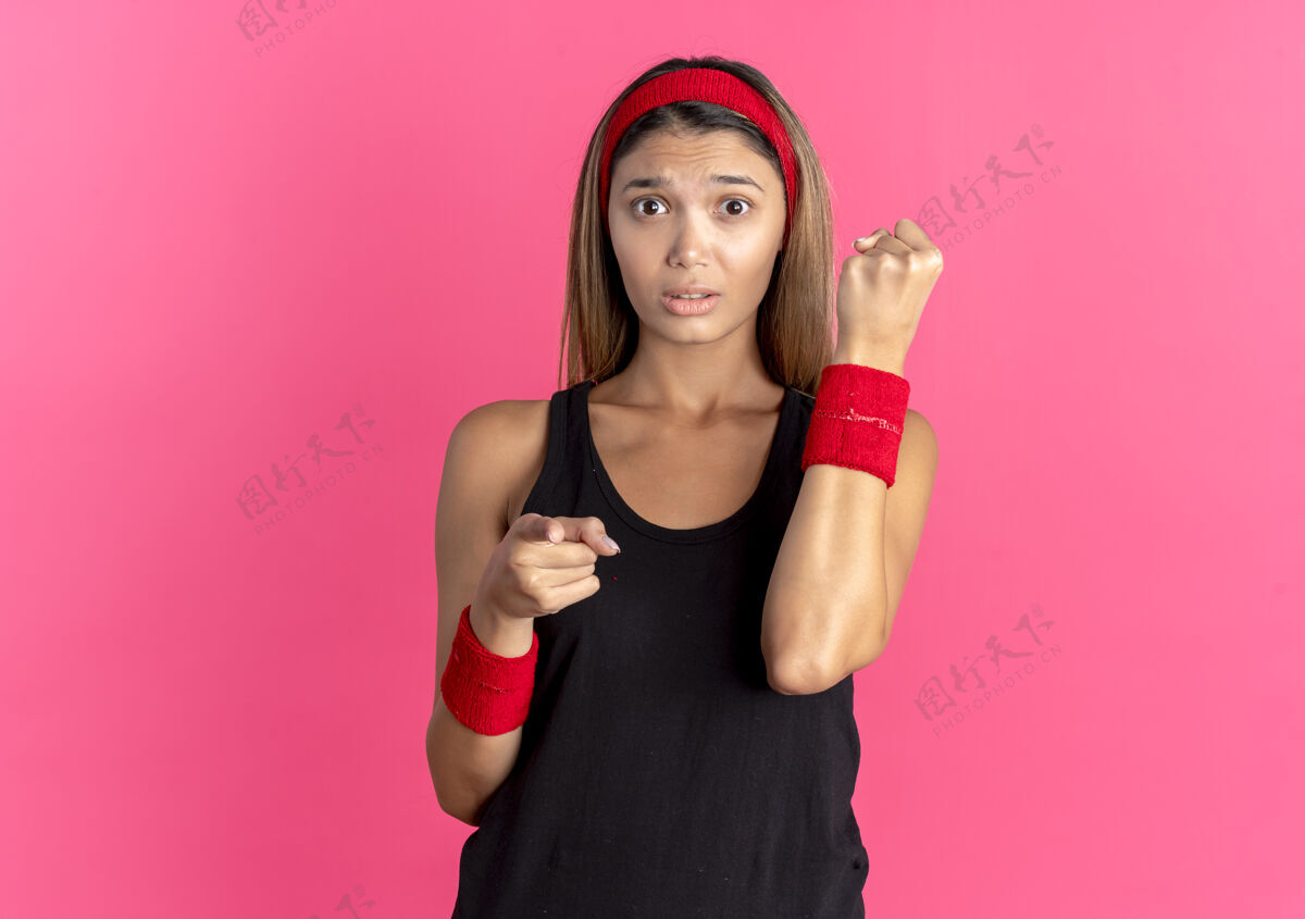 粉色身穿黑色运动服 头戴红色头带的年轻健身女孩看起来很困惑 握紧拳头 用手指指着粉色的相机年轻握紧困惑