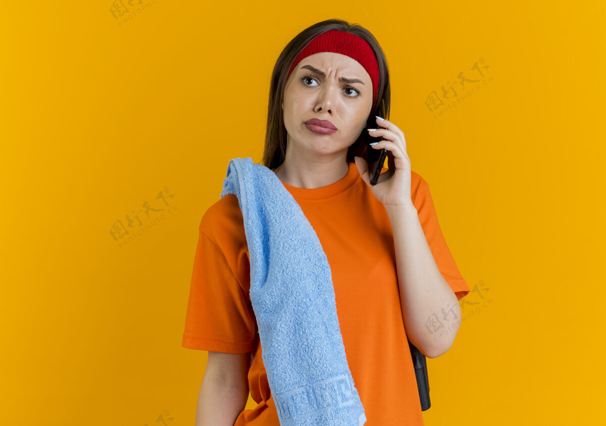 毛巾皱眉的年轻运动女性戴着头带和腕带 肩上扛着跳绳和毛巾 一边打电话一边看穿橙色说话