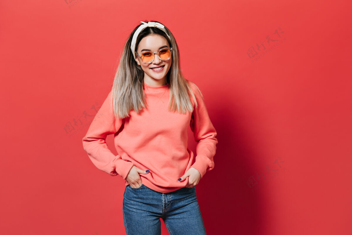 美丽戴着发带 戴着橙色眼镜和运动衫 在红墙上摆姿势的女人毛衣兴奋时尚