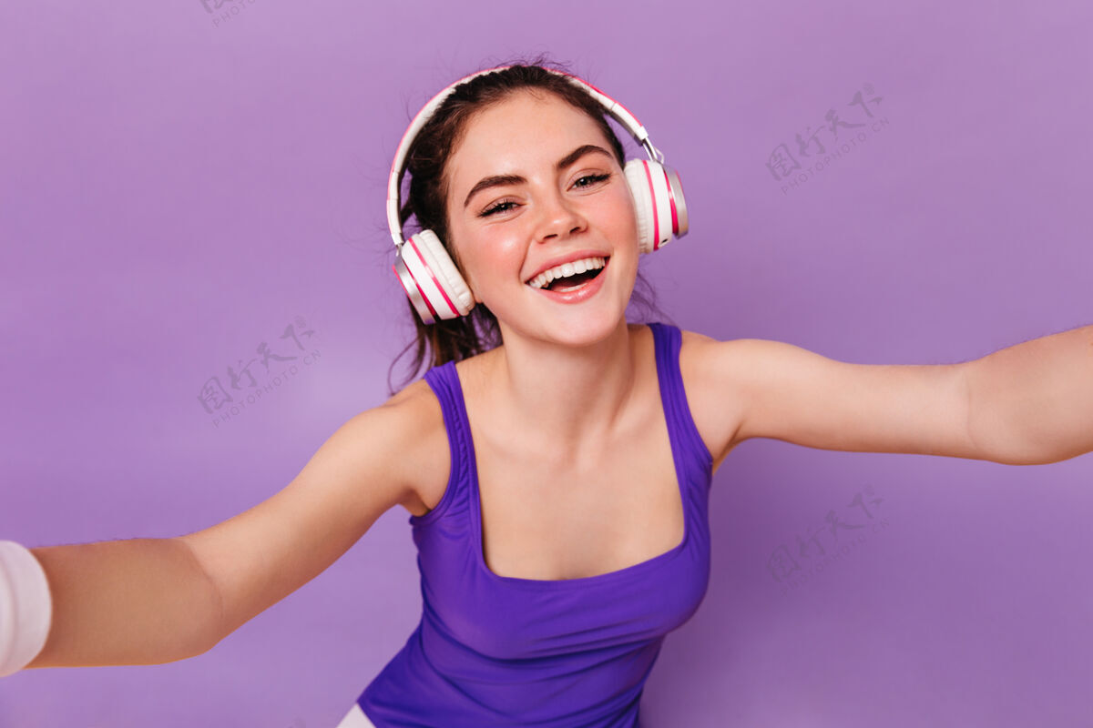 音乐穿着健身上衣戴着耳机在紫色墙上自拍的快乐女人的特写肖像时尚姿势漂亮