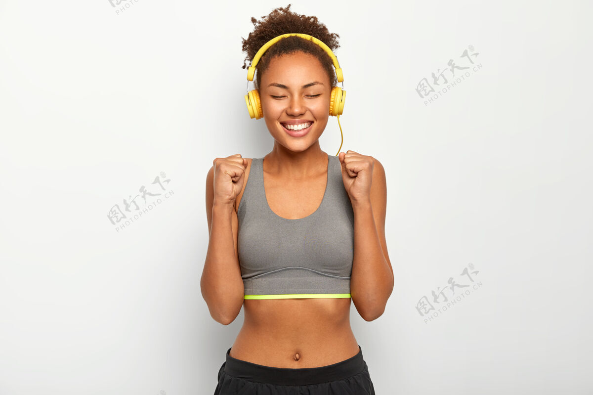 训练快乐的女人举起紧握的拳头 庆祝胜利的比赛 穿着运动内衣 通过耳机听音乐高兴民族运动员