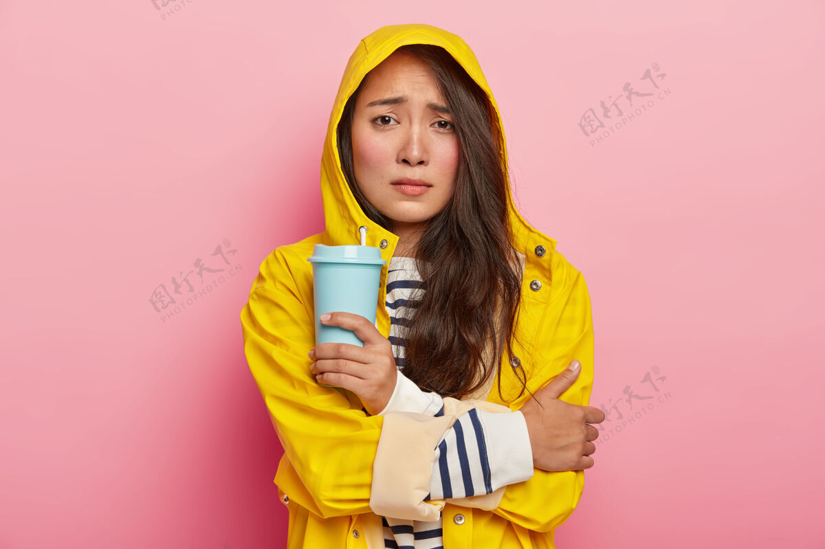 毛衣不满意的亚洲女人的照片笑脸 双臂交叉 在雨中行走后因寒冷而颤抖 穿着黄色雨衣 拿着外卖咖啡 用热饮料取暖皱眉脸不喜欢