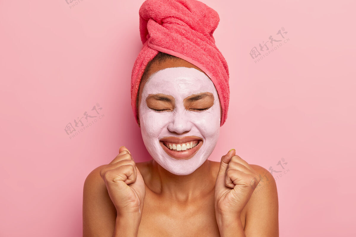 非洲充满活力的快乐女人脸上戴着泥面具 头发上裹着毛巾 笑容灿烂 从快乐中握紧拳头 隔着粉色的墙壁头像高兴沐浴