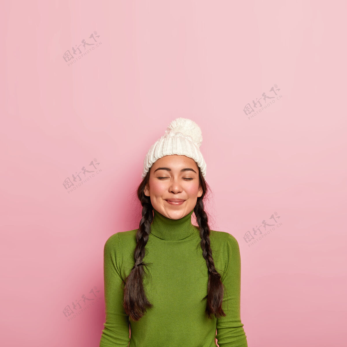 冬天高兴的黑发女孩闭上眼睛 想着和朋友愉快的见面 戴着白帽子和绿色高领毛衣 靠着粉色的墙站着胭脂衣服人