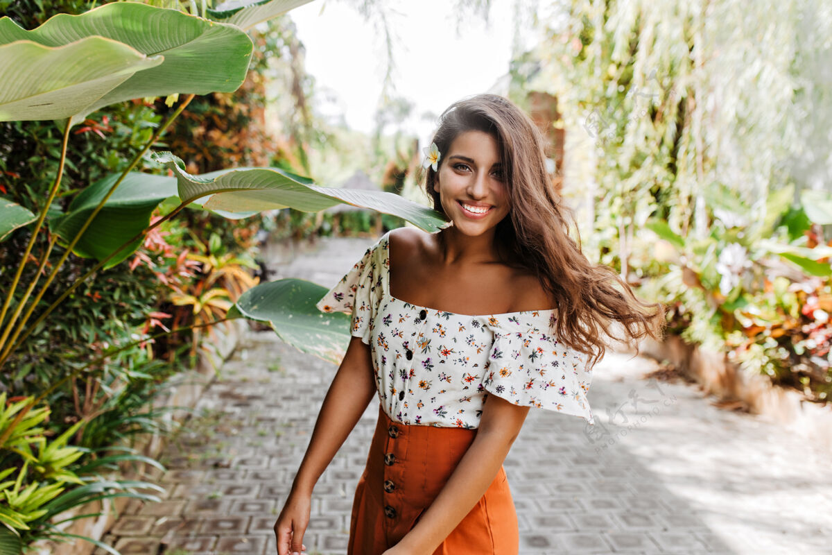 快乐一位穿着夏装 皮肤黝黑的可爱女士在热带植物的墙上真诚地微笑着肖像微笑女人