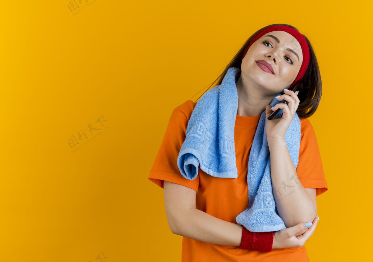 看高兴的年轻运动妇女戴着头带和手环与毛巾围着脖子看一边把手放在手肘上讲电话周围年轻放