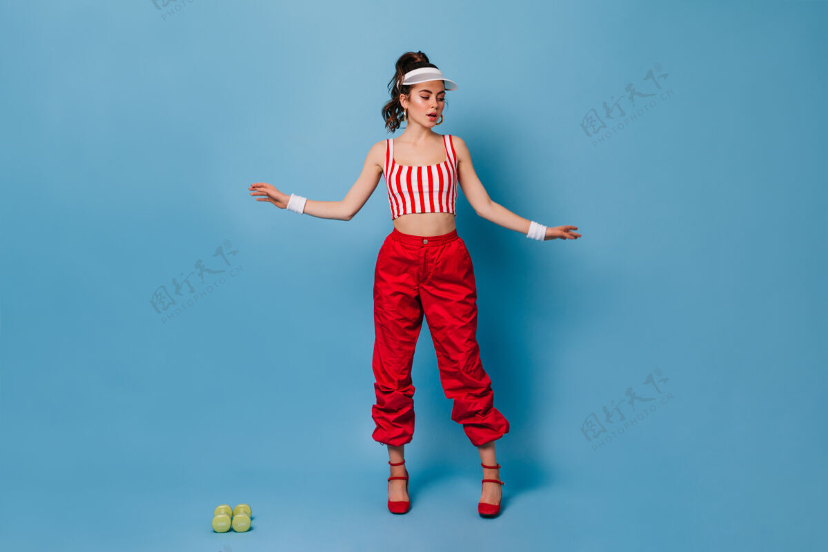 哑铃穿着80年代运动风格的时髦服装的女人在蓝色墙上摆姿势漂亮高跟鞋优雅